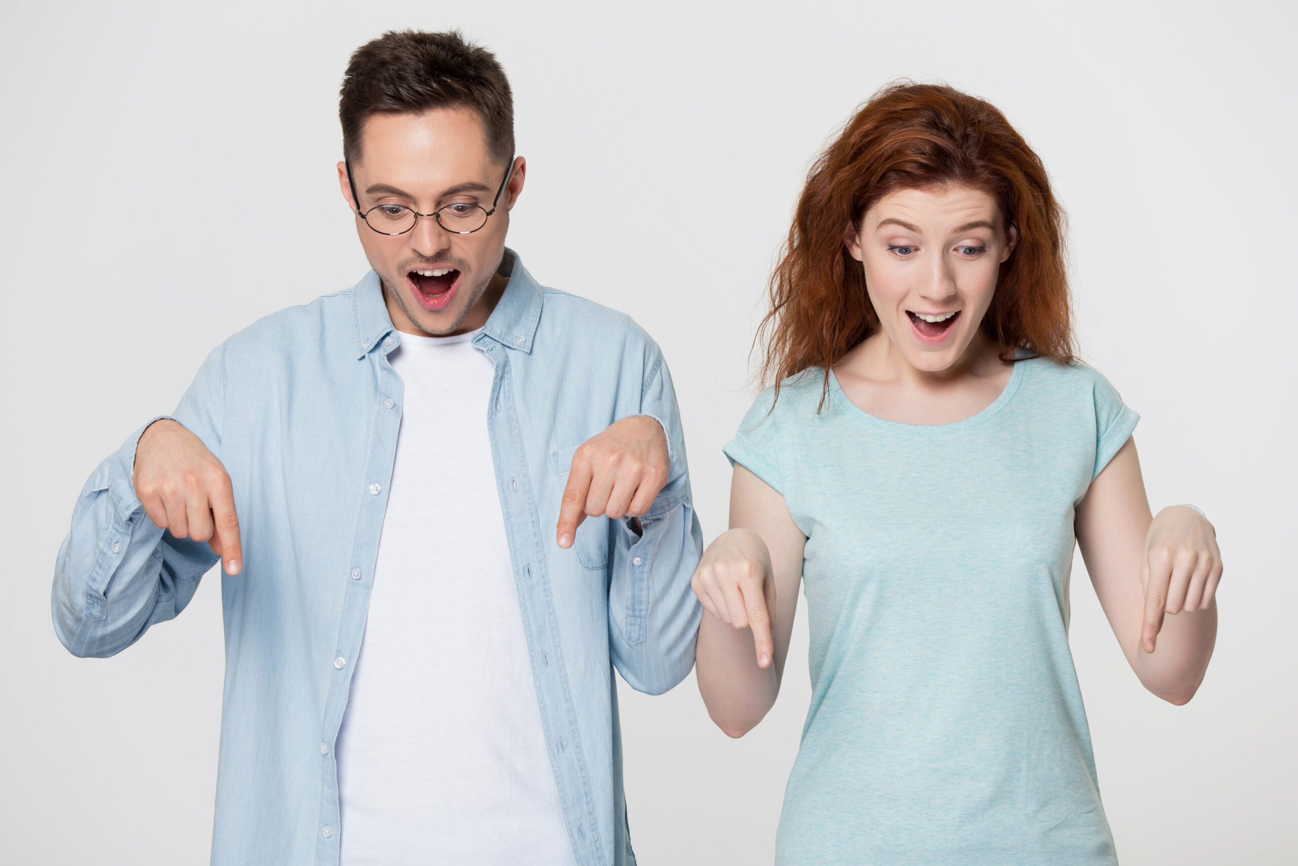 Young couple feels amazed point fingers looking down pose over grey, redhead girl guy in glasses advertise something unbelievable recommend new store trendy shop, subscribe our channel gesture concept
